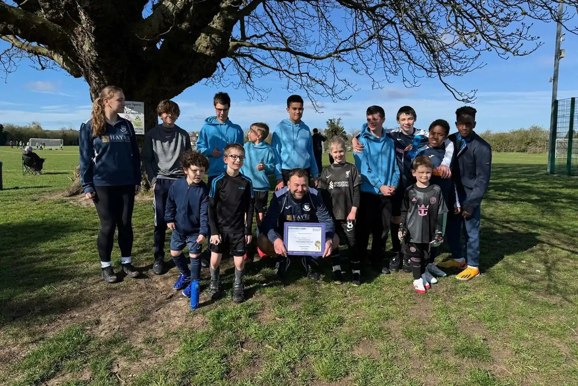 Young football players and coaches celebrating outdoors under a large tree after winning a match.